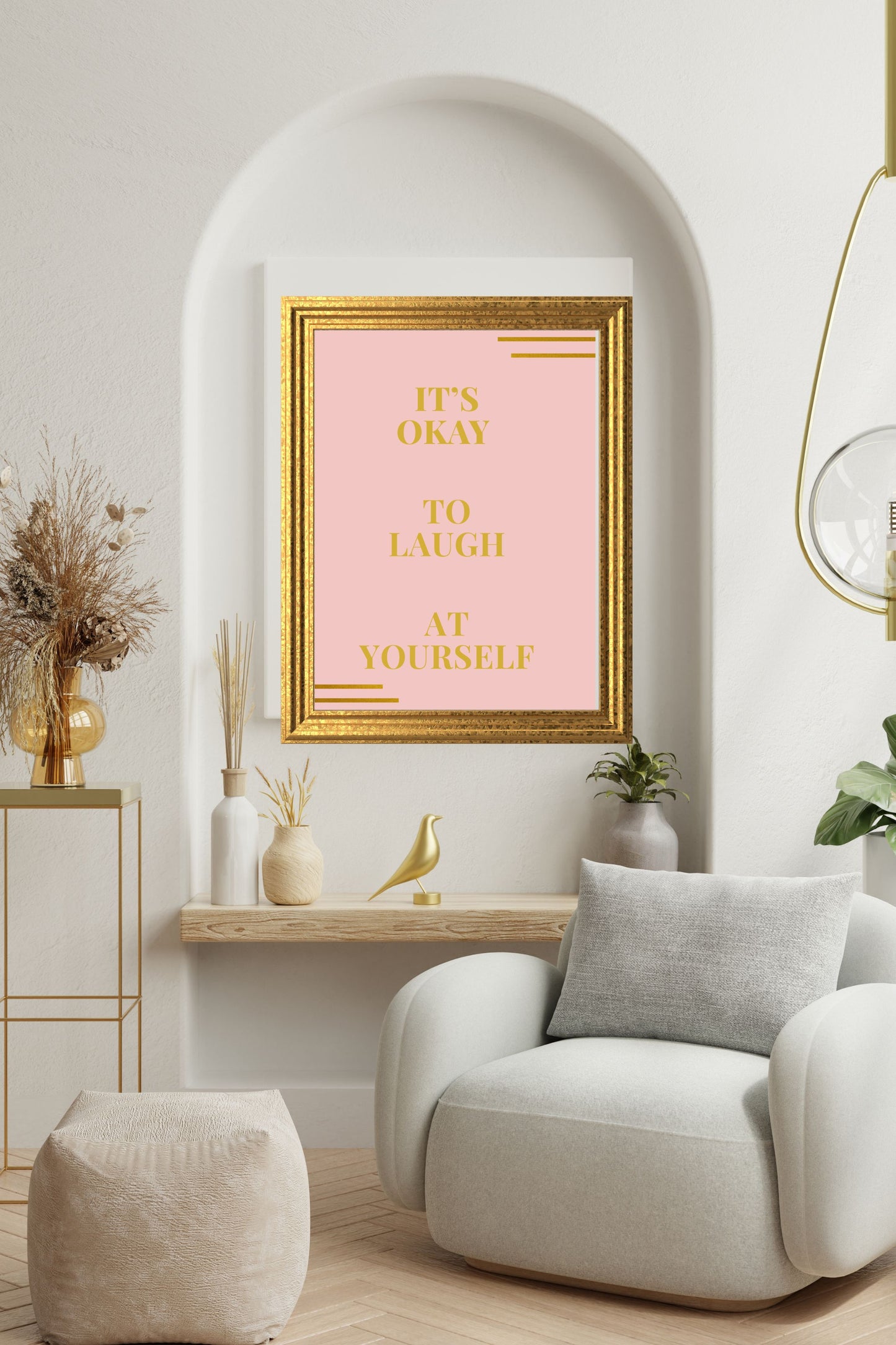 Motivational printable wall art featuring the quote "It’s Okay to Laugh at Yourself" in gold on a blush background, framed in gold and displayed in a modern white alcove above a plush armchair, with neutral decor and dried floral accents.