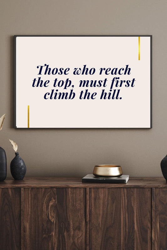 Motivational printable wall art featuring the quote "Those who reach the top, must first climb the hill" in navy serif font with gold accent lines, styled above a walnut sideboard with gold bowl, black vases, and a warm neutral wall—creating a refined, inspiring atmosphere.