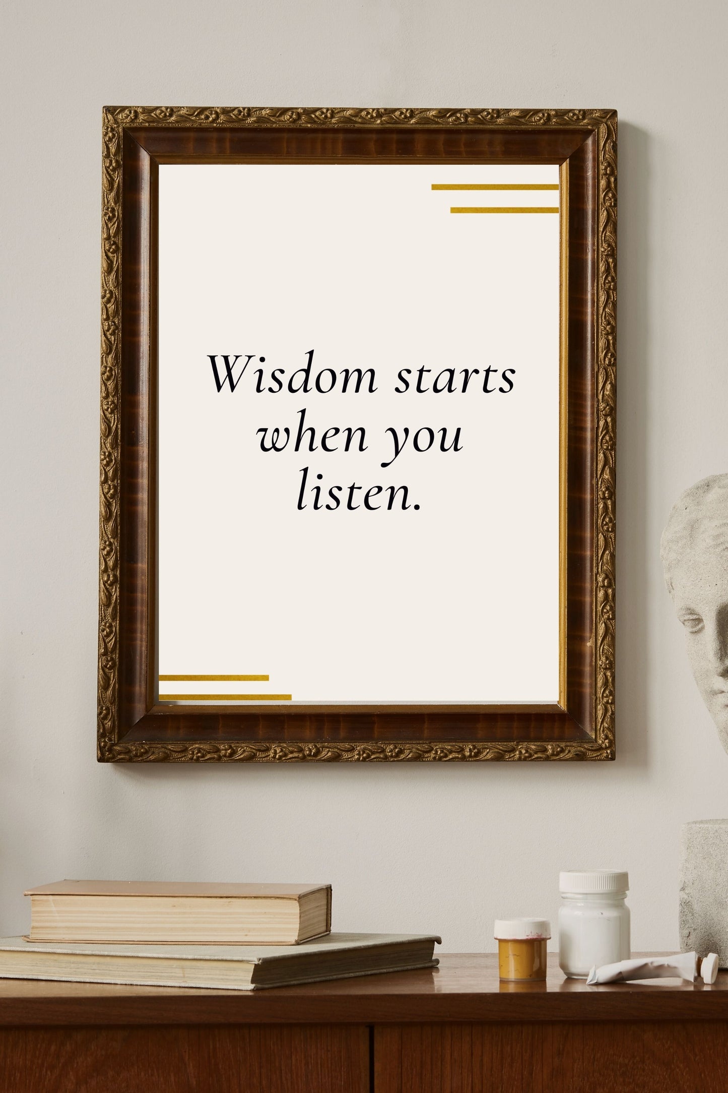 Motivational printable wall art featuring the quote "Wisdom starts when you listen" in elegant black script with gold minimalist accents, framed in ornate brown-gold, styled above books and creative supplies in a serene study setting.